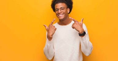 Young african american man over an orange wall smiles, pointing mouth