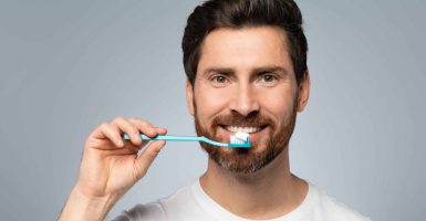 Morning procedure, oral care and perfect smile. Bearded man in white t-shirt holding toothbrush with paste and smiling at camera over grey studio background