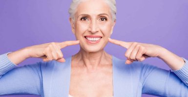 Concept of having strong healthy white perfect teeth at old age. Portrait of old lady with beaming smile pointing on her teeth, isolated over violet background