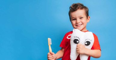 A smiling boy with healthy teeth holds a plush tooth and a toothbrush on a blue isolated background. Oral hygiene. Pediatric dentistry. Prevention of caries. A place for your text