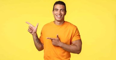 Cheerful modern man with tattooed arm, pointing left as promoting online store, invite guest join party, smiling joyfully, recommend try something, give advice what place visit, yellow background.