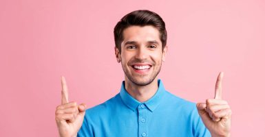 Photo of cute brunet millennial guy point up empty space wear blue t-shirt isolated on pink color background.